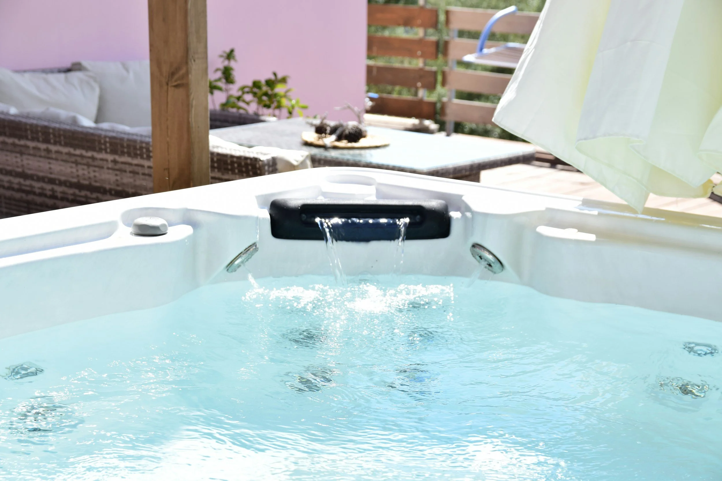 A hot tub with bubbling water in an outdoor patio area, surrounded by a wooden fence and a pink wall, with a dining table and chairs in the background and a towel hanging on a rail.