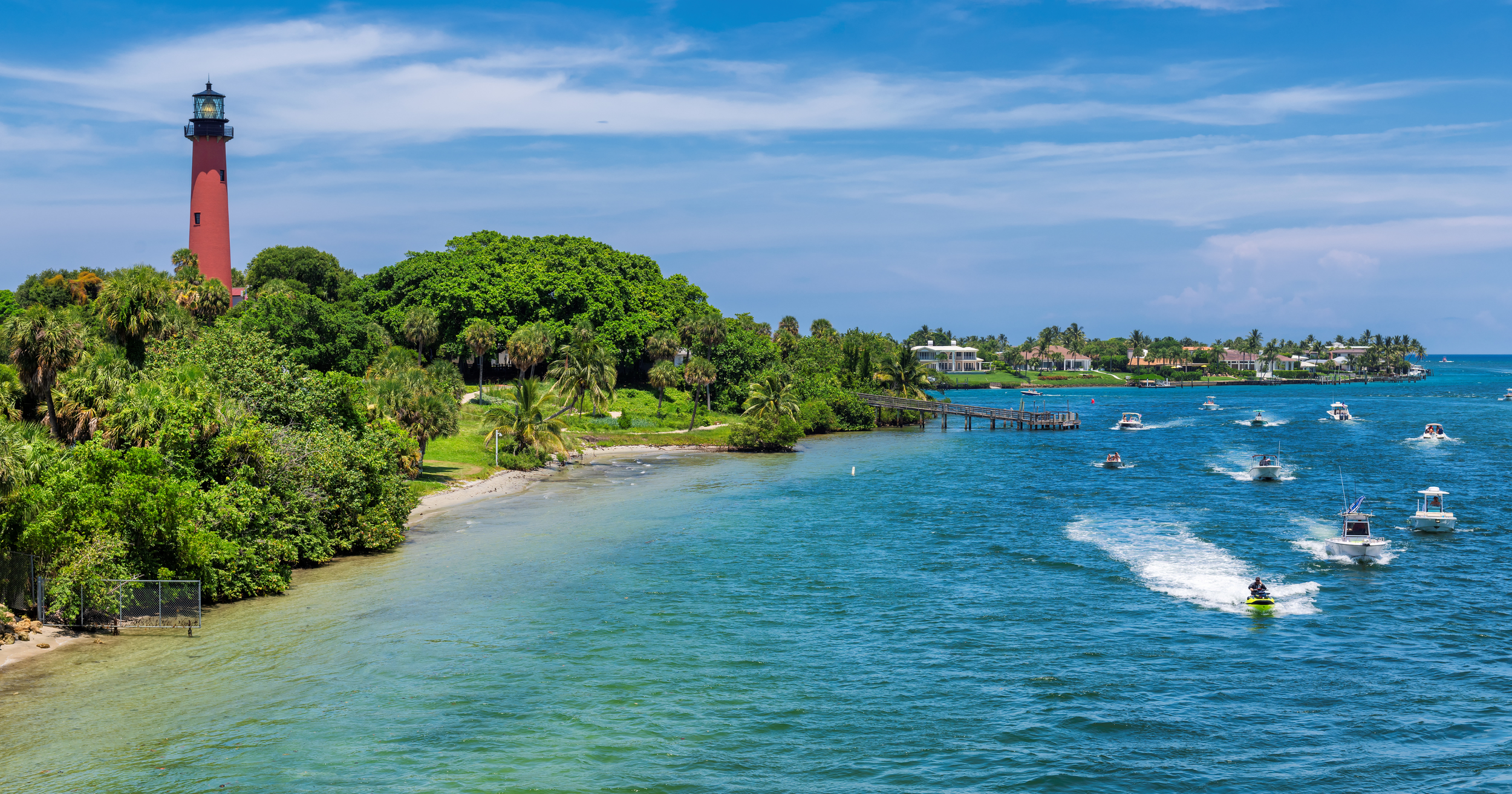 A coastal scene with a lighthouse on the left, lush greenery, a sandy beach, and several boats on the water near a pier, under a partly cloudy sky.