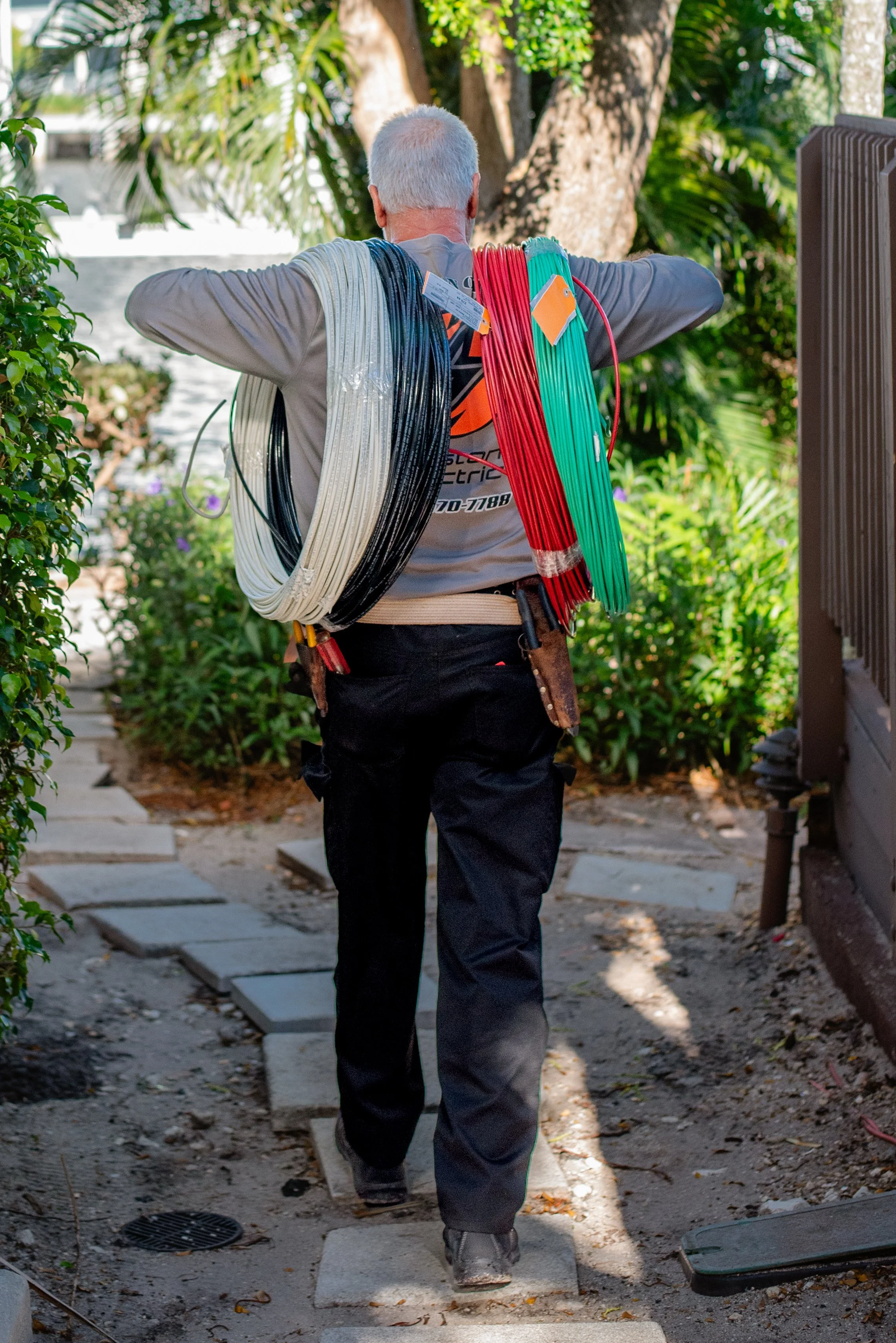 A worker with gray hair walking on stones on a garden pathway, carrying colorful wires or cables over his shoulders, wearing a utility belt and dark pants, with lush greenery in the background.