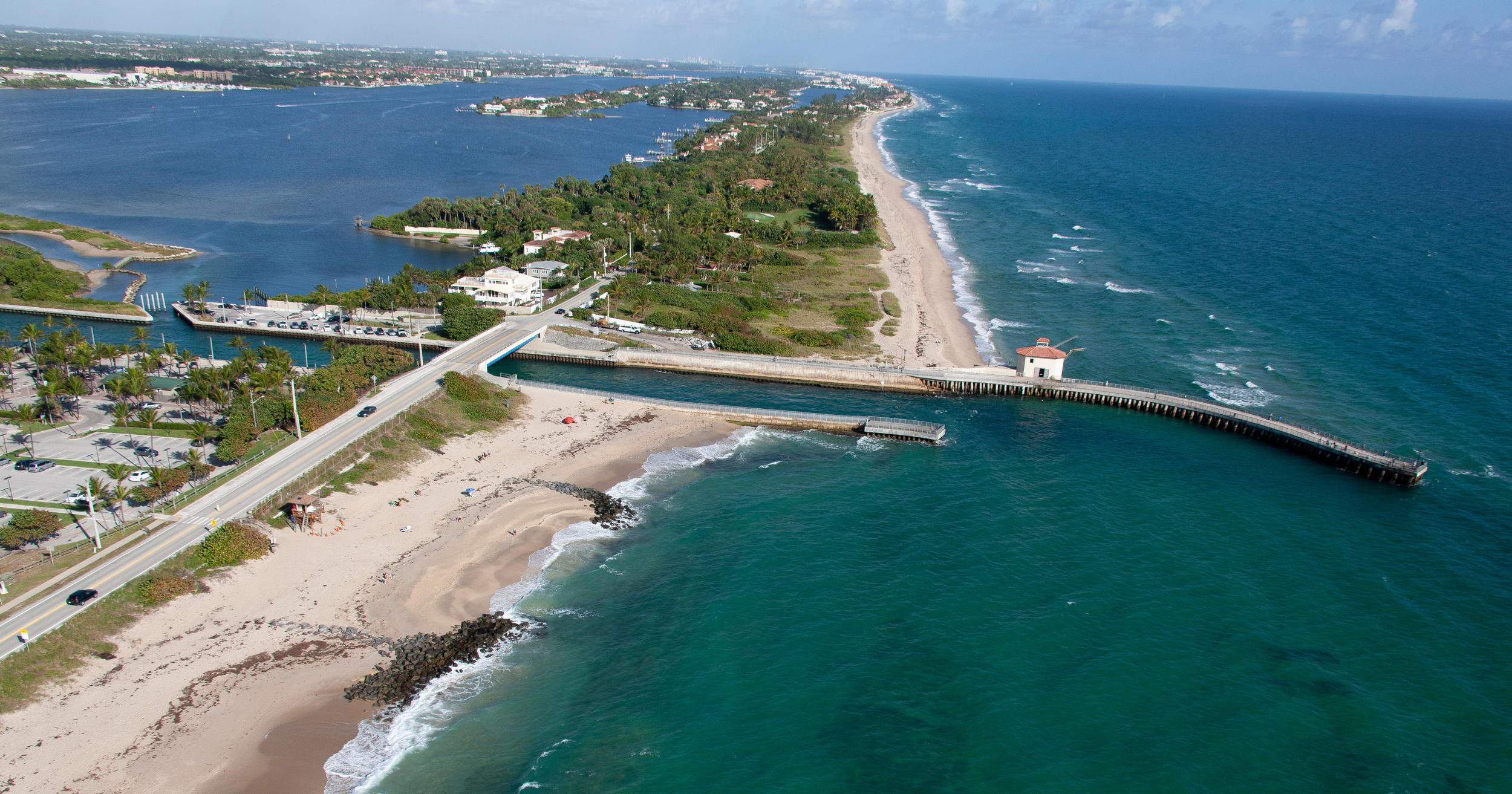 Aerial view of a coastal area featuring a bridge along a sandy beach, a small white building at the end of the pier, surrounding greenery, and a body of water with boats, set against a blue sky with clouds.