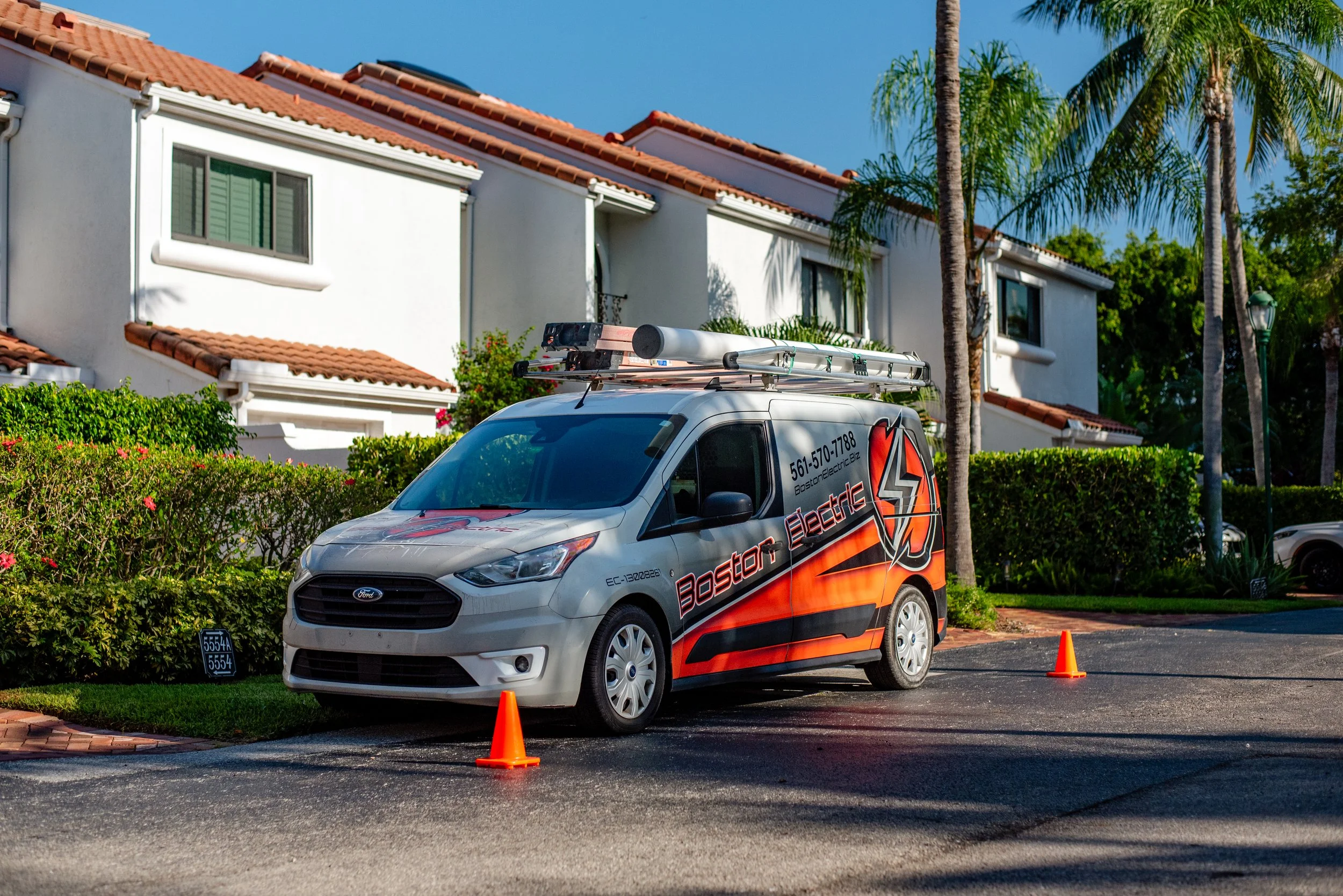 A white service van parked on the street with orange traffic cones in front. The van has the logo 'Boston Electric' and a phone number on the side. Behind the van, there is a white multi-story residential building with a red tile roof and large windows. Palm trees and bushes are in the background under a clear blue sky.