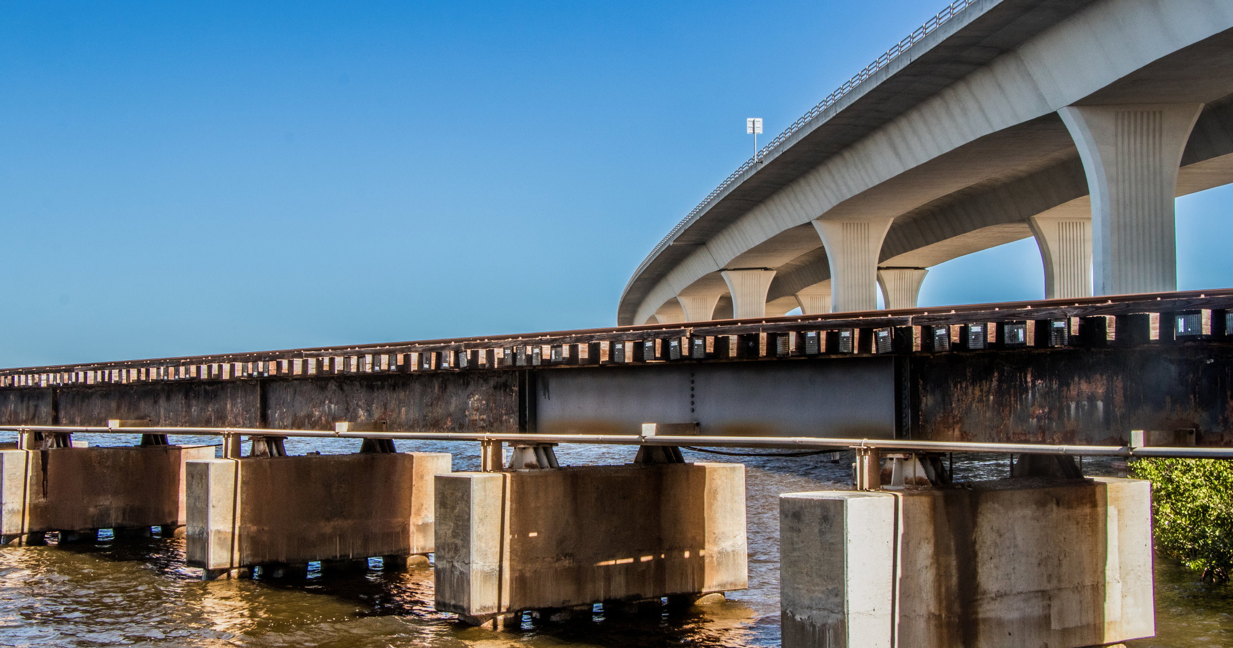 A large concrete bridge extending over a body of water, supported by multiple large columns, with a clear blue sky in the background.