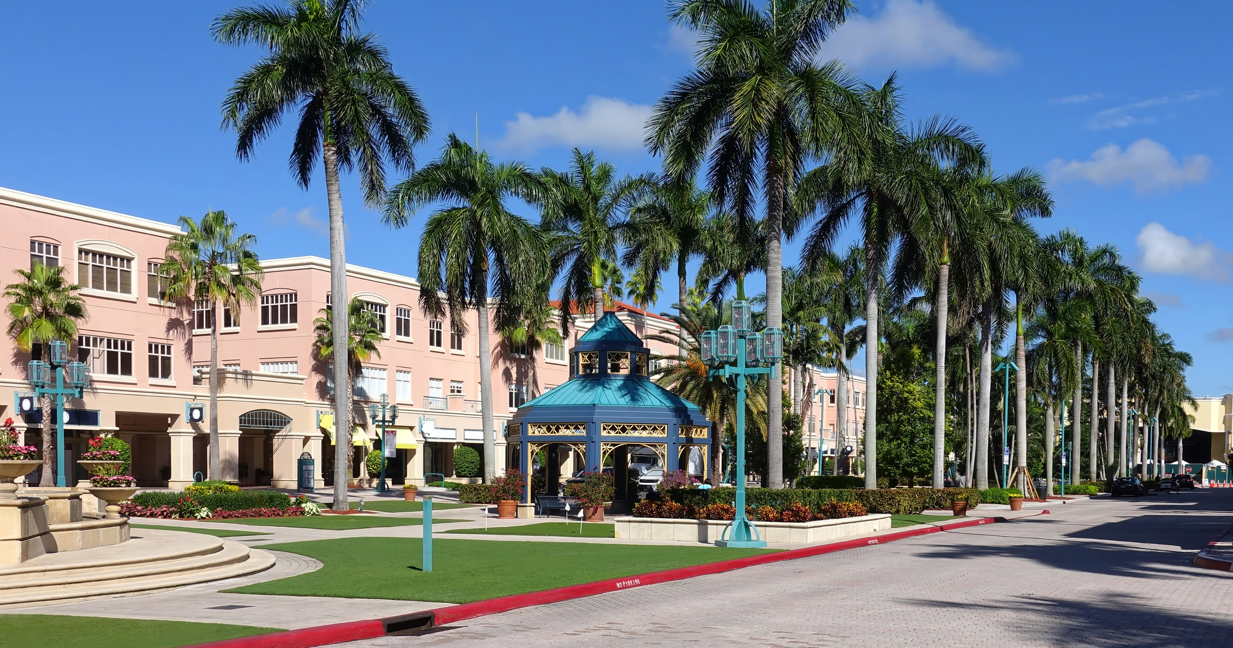 A street scene with pink buildings, tall palm trees, decorative lampposts, and a gazebo with a blue roof under a clear blue sky.