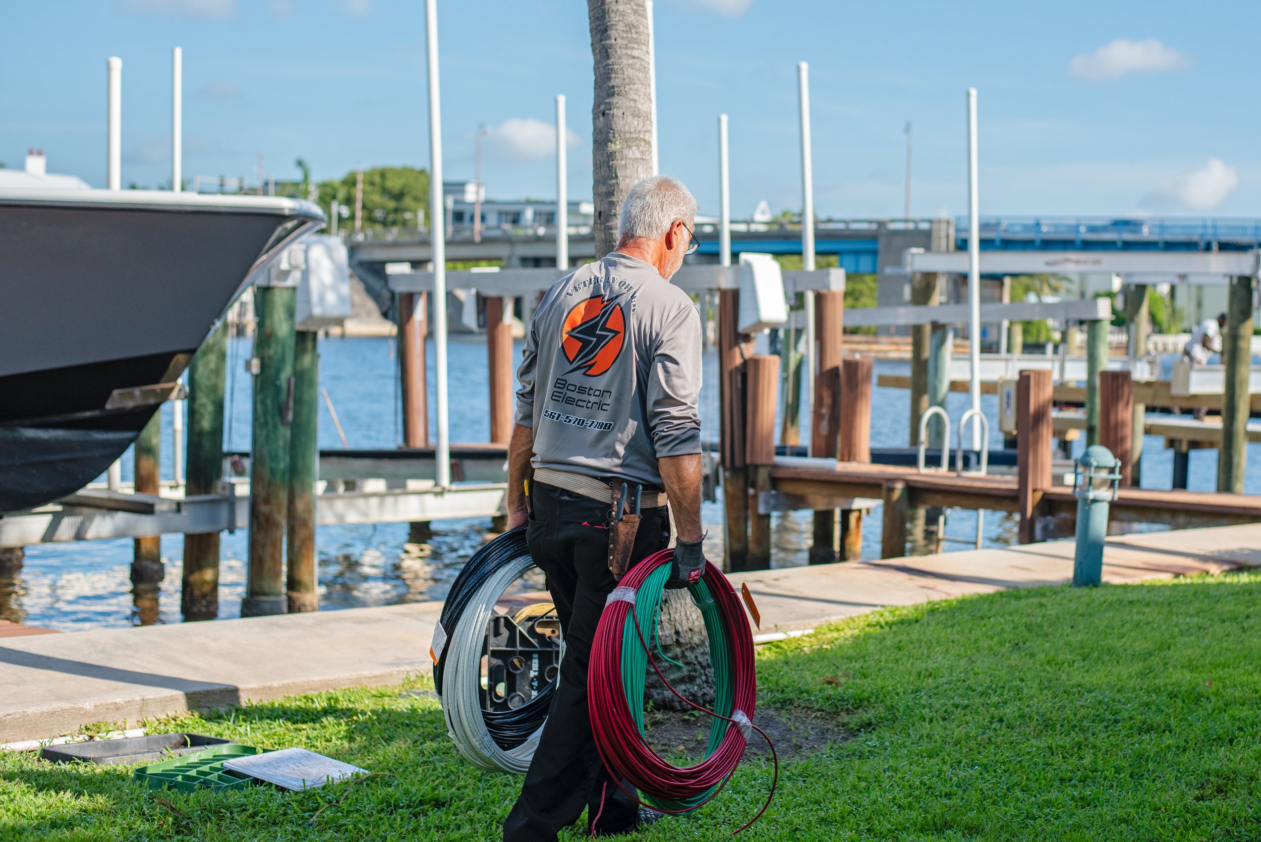 An older man with gray hair and glasses is carrying electrical cables and equipment at a marina dock, wearing a gray shirt with a Boston Electric logo on the back.