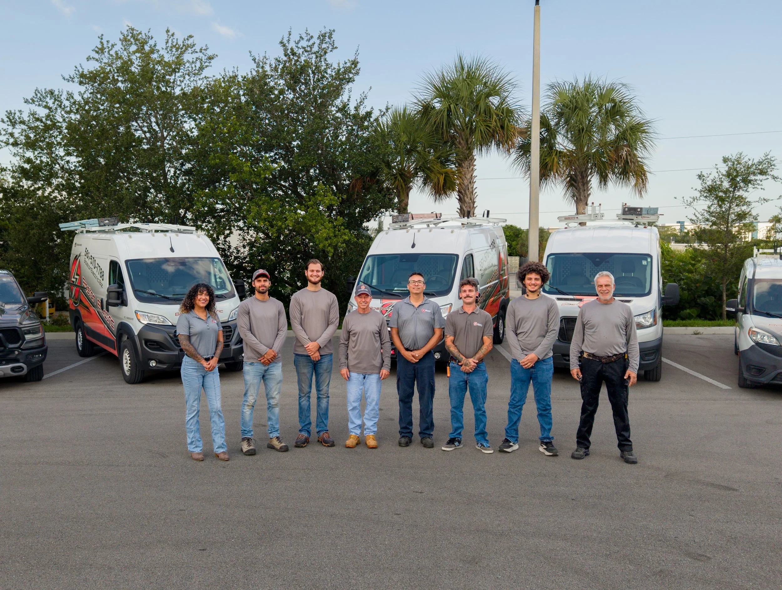 A group of nine people standing in a parking lot in front of three white service vans. The background includes trees, a utility pole, and a clear blue sky.