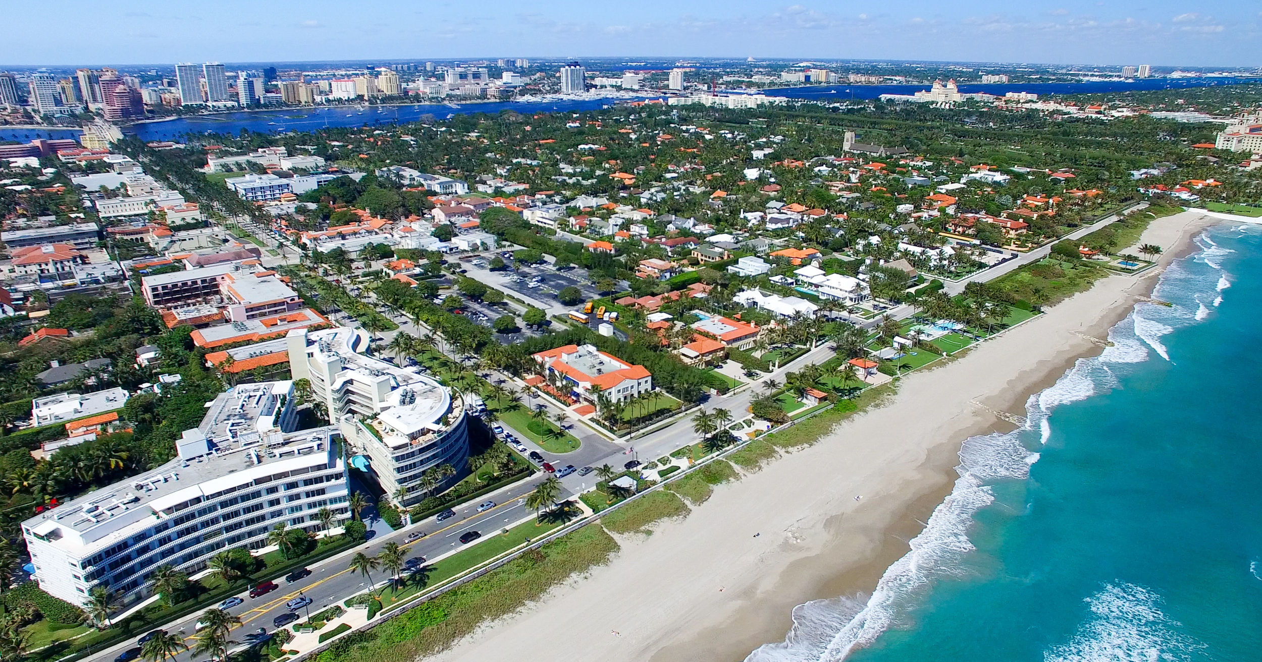 Aerial view of a coastal city showing a sandy beach along the ocean, residential houses with red-tiled roofs, a downtown area with tall buildings and skyscrapers, and a river running through the city with bridges spanning it.