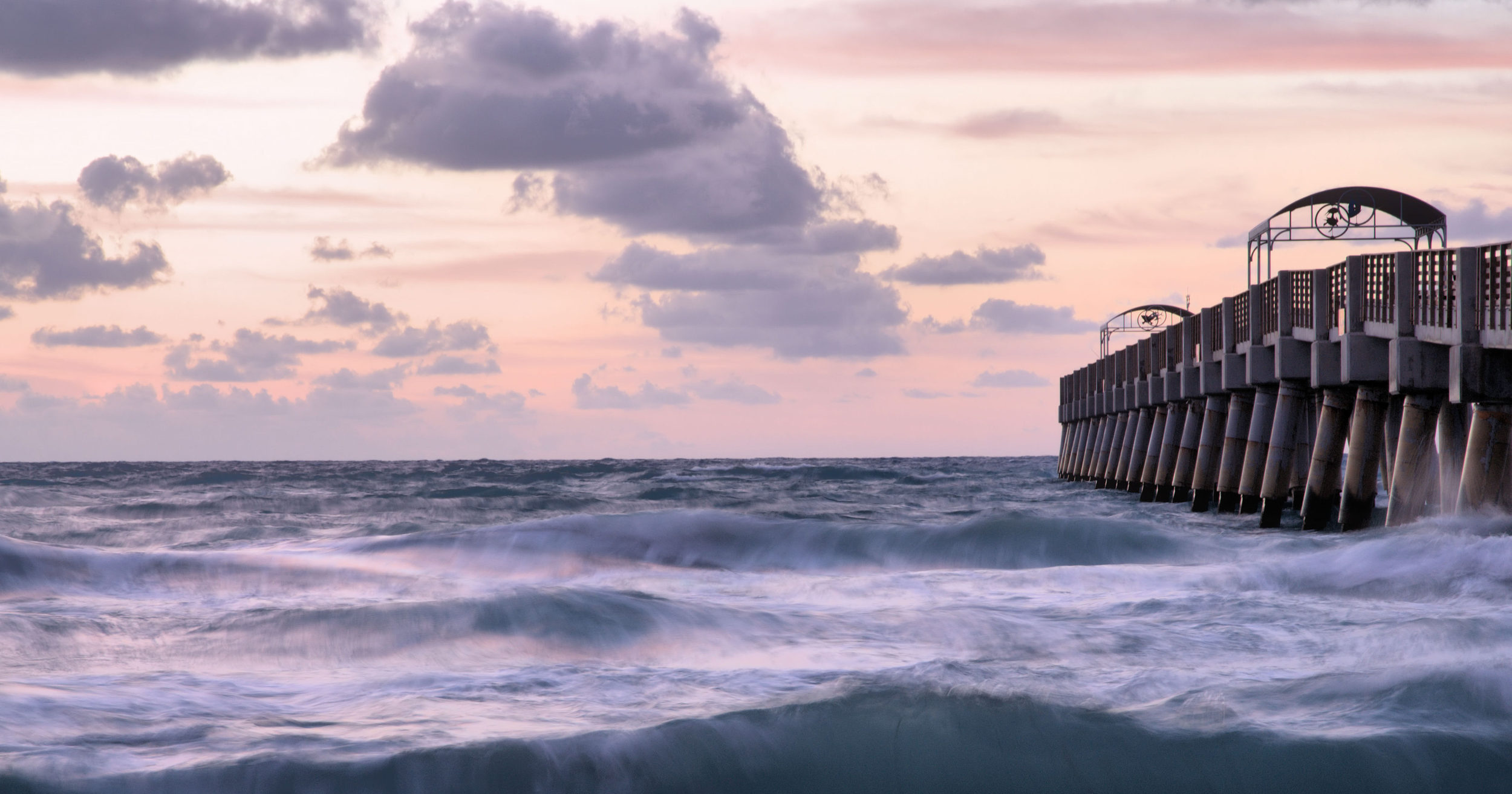 A pier extending into the ocean at sunset with waves crashing against the structure, and a sky filled with pink, purple, and grey clouds.
