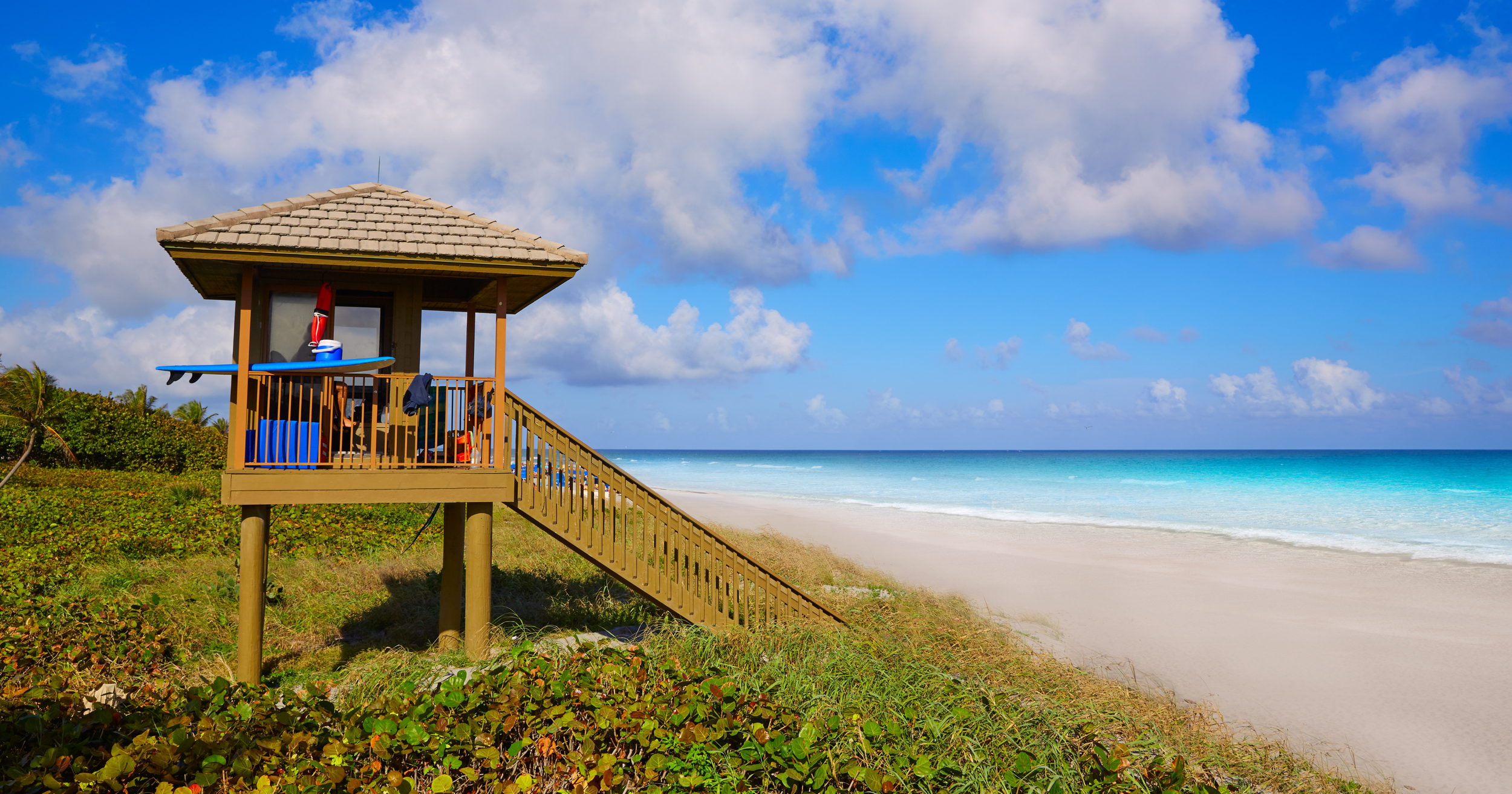 A wooden lifeguard tower with a sandy pathway leading up to it overlooking a beach and ocean under a blue sky with white clouds.