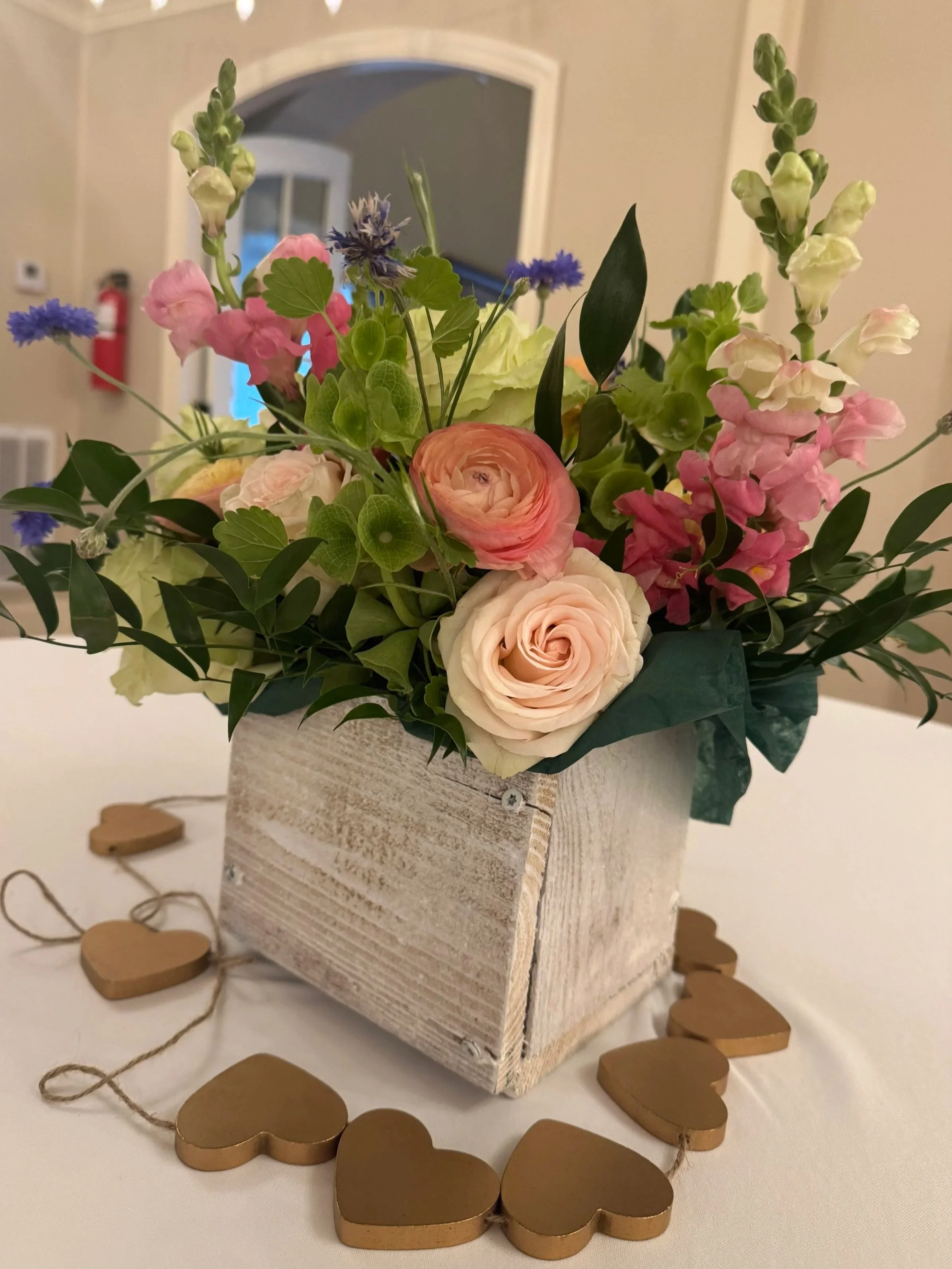 A bouquet of mixed flowers in a white wooden box surrounded by a string of wooden hearts on a white surface.