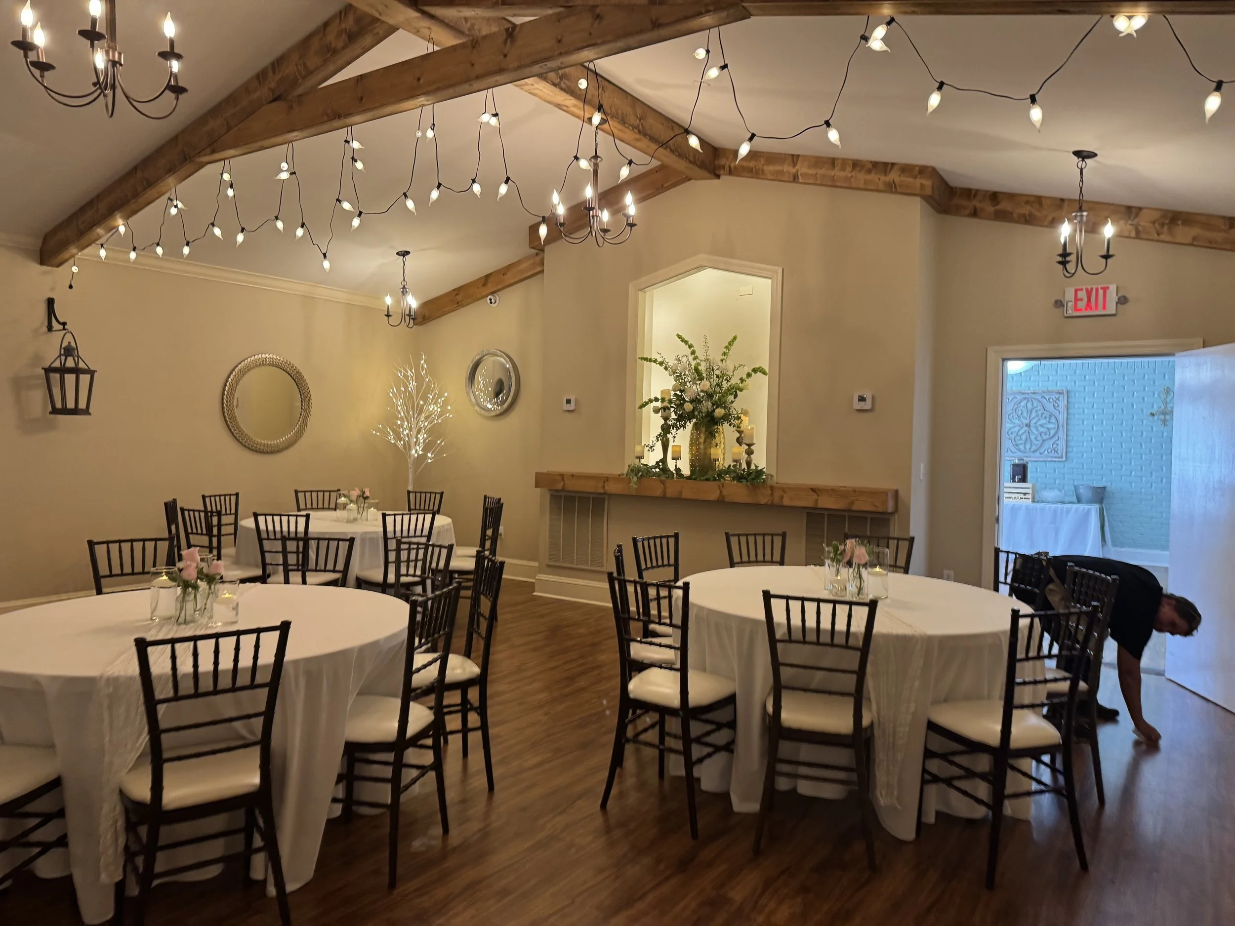 A decorated event space with round tables covered in white tablecloths, black chairs with white cushions, floral centerpieces, and string lights hanging from wooden beams. There are also chandeliers and wall lanterns, with a doorway leading to another room.