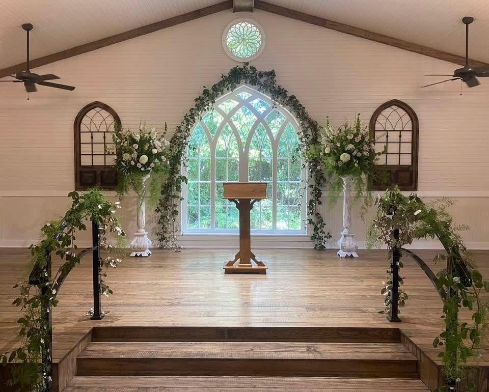 Wedding ceremony setup with a wooden podium on a raised platform, decorated with greenery and white flowers. Behind it is a large arched window with sunlight streaming in, flanked by two window frames with plants. The room has a vaulted ceiling with two ceiling fans and a small round decorative window.