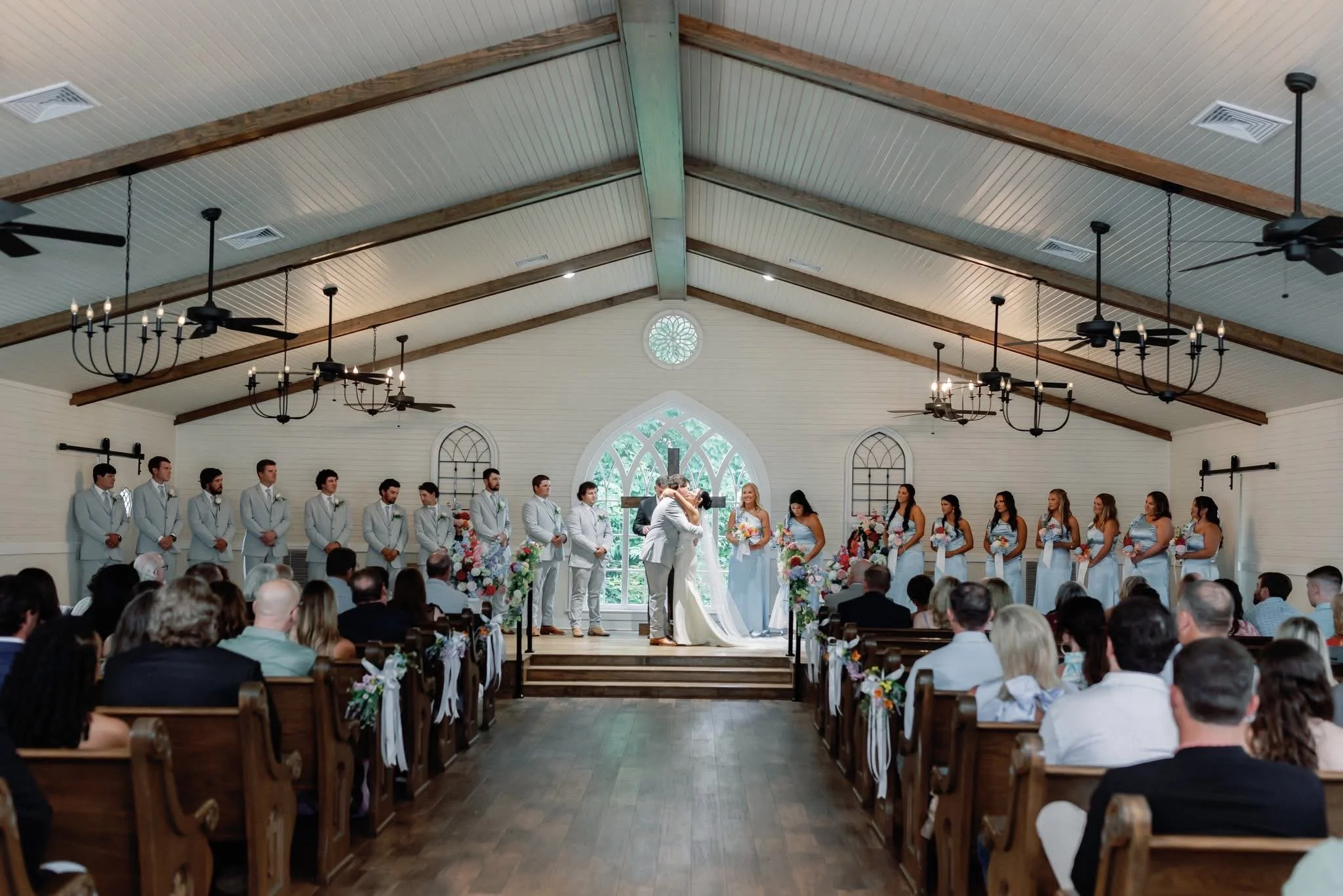 A church with a wooden beam ceiling and a bride and groom are exchanging vows at the altar, surrounded by bridesmaids and groomsmen. Guests are seated facing the ceremony. Photography JB Photography