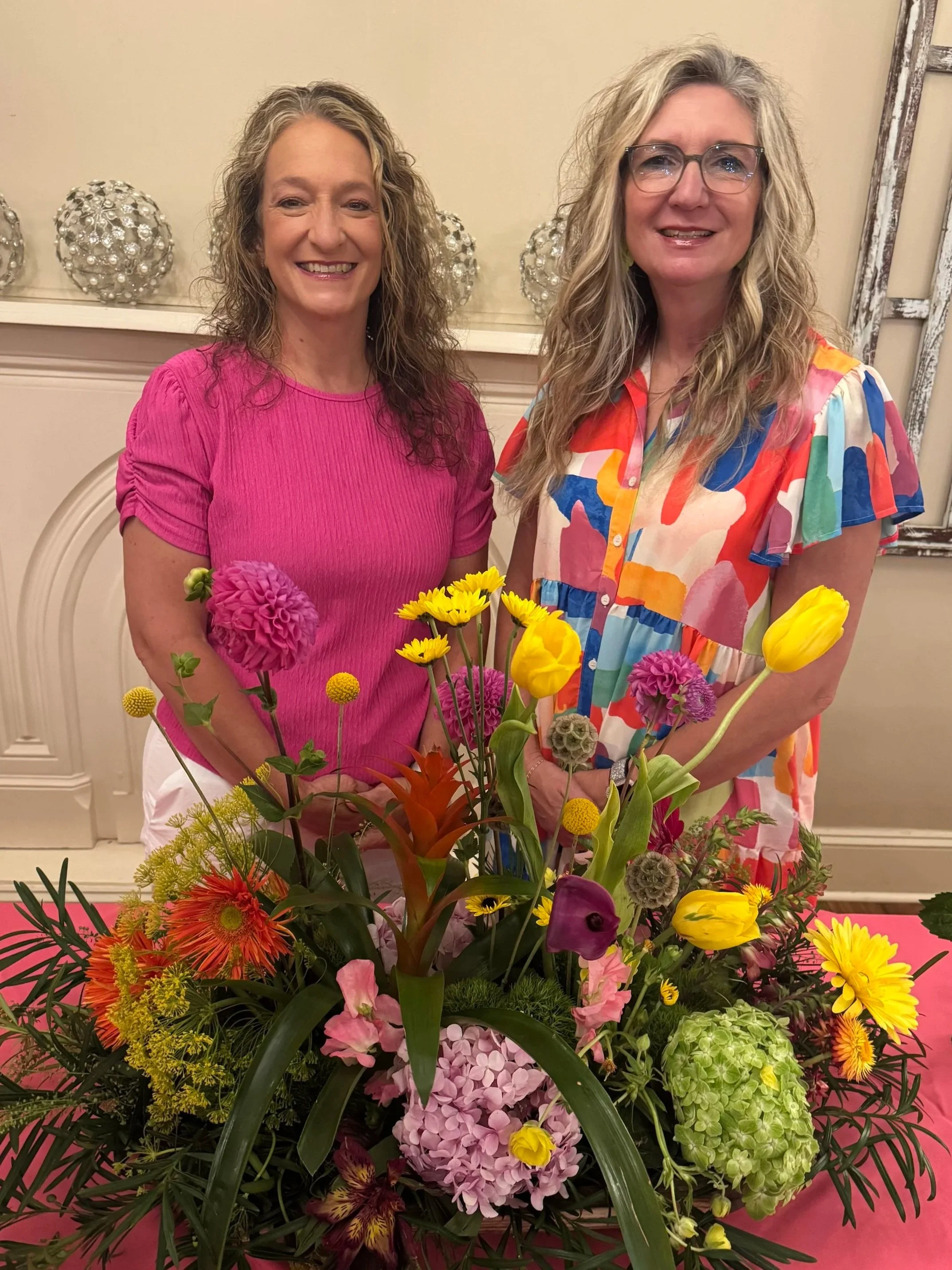 Two women smiling behind a large colorful bouquet of flowers in a room with a beige wall and decorative spheres on a shelf.