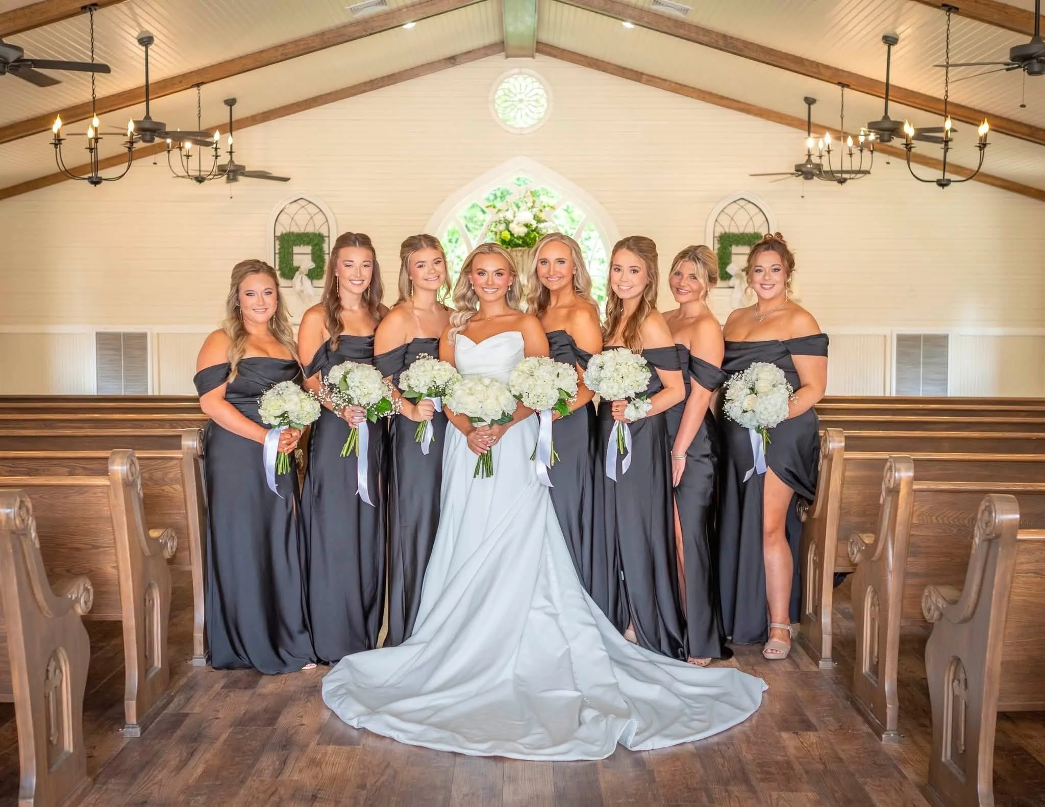 Inside a church with a high ceiling, chandeliers, and fans, seven women dressed in black bridesmaid dresses and a bride in a white wedding gown holding bouquets, standing together for a photo.