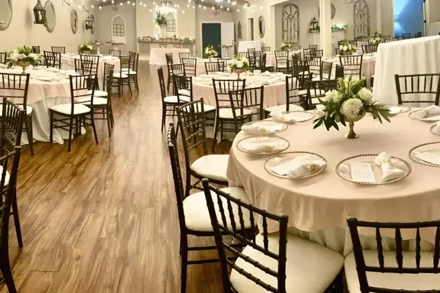 Elegantly decorated banquet hall with round tables covered in white tablecloths, each with a floral centerpiece, set for a formal event, with wooden chairs arranged around the tables and soft lighting.