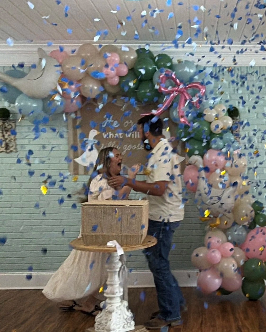 Two children celebrating with colorful balloons and confetti, one girl with her mouth open in excitement, woman handing her a gift box, decorated indoor setting with balloons and wall decor.