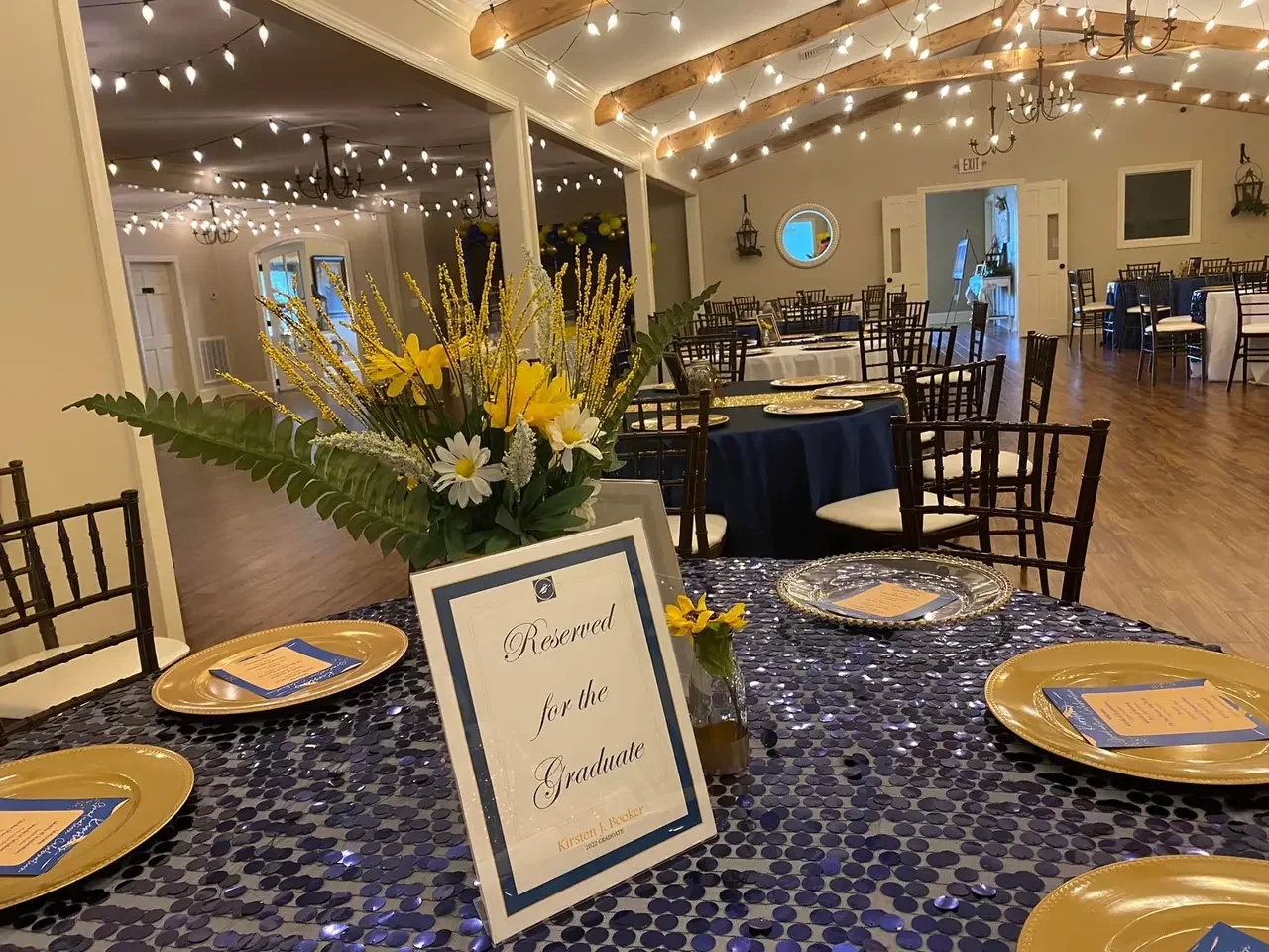 Decorated banquet hall with round tables covered in dark tablecloths, gold plates, and reserved signs for a graduation celebration. The room features string lights, chandeliers, and flowers with yellow and white blooms and green foliage.