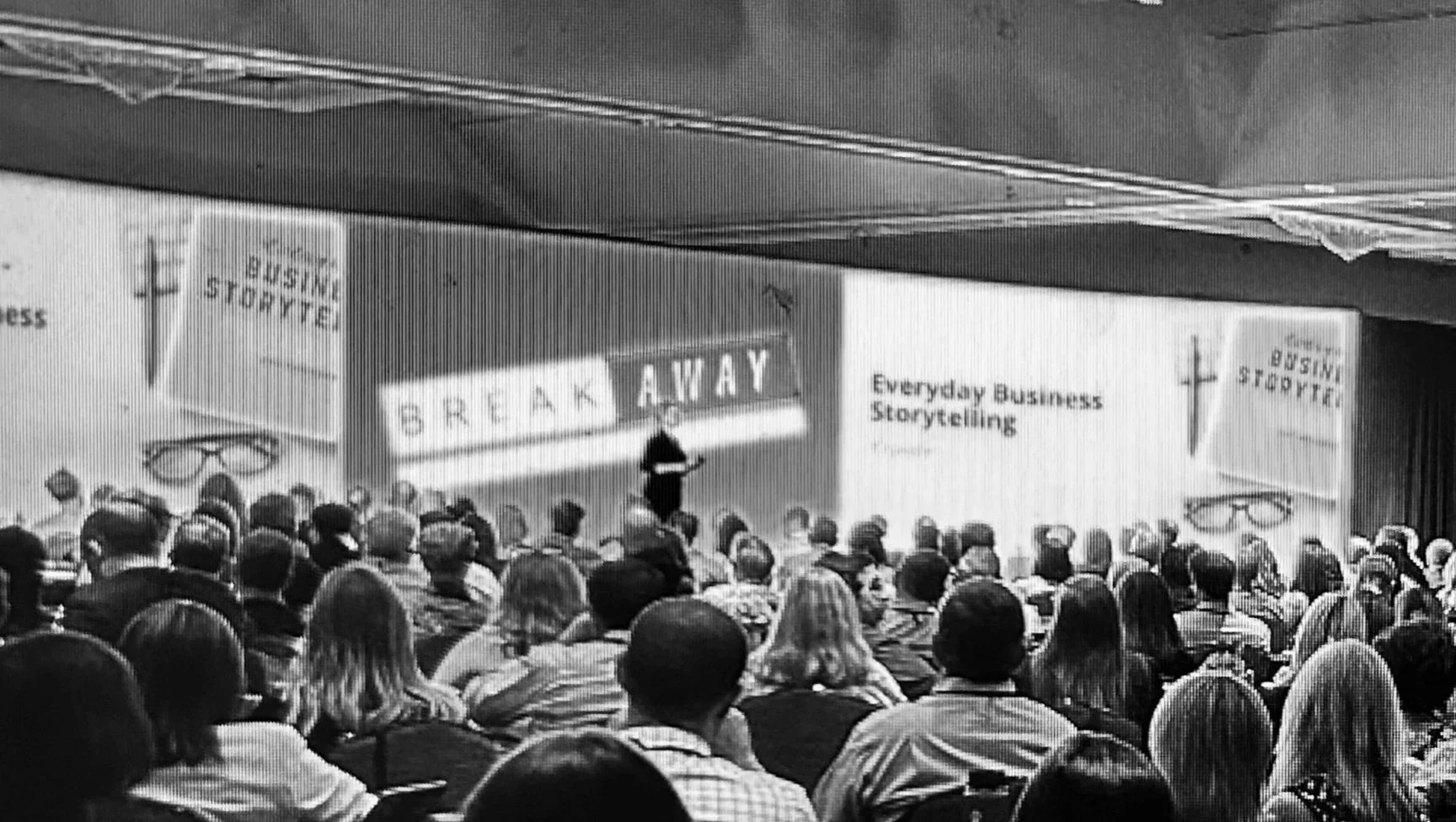 A black and white photo shows an audience attending a presentation on everyday business storytelling. The audience is facing a stage with a speaker standing in front of large screens displaying the words 'SPEAK AWAY' and 'Everyday Business Storytelling.' The crowd appears to be engaged, and the setting seems to be a conference or seminar.
