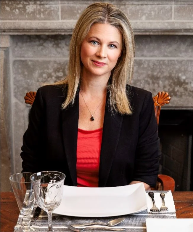 A woman with blonde hair sitting at a dining table, smiling, with a gray stone fireplace in the background, wearing a black blazer over a red top.