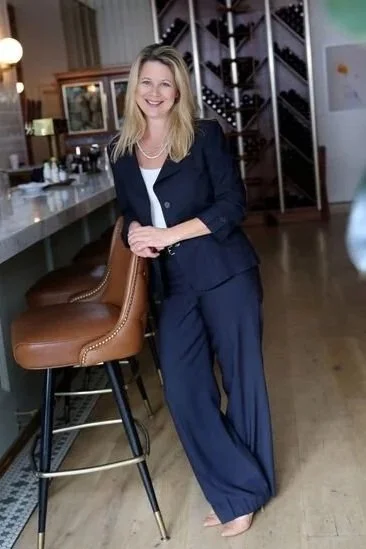 A smiling woman in a navy blue pantsuit standing next to a bar counter with brown leather stools in a cozy, well-lit room.
