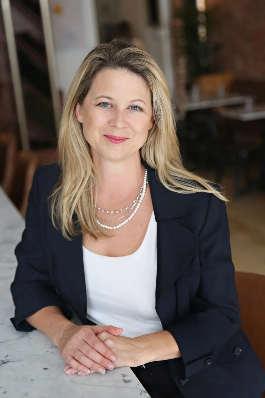 A woman with blonde hair and blue eyes, wearing a black blazer over a white top and layered pearl necklaces, sitting at a marble table in a warmly lit cafe or restaurant.
