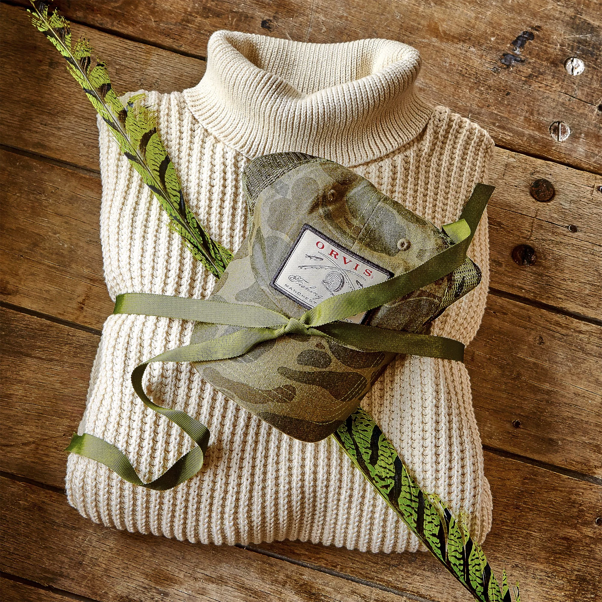 A folded beige knitted sweater with a turtleneck is placed on a wooden surface. There are green and black patterned leaves, possibly artificial, arranged on top of the sweater. A partially visible tag or packaging with some text is also seen.