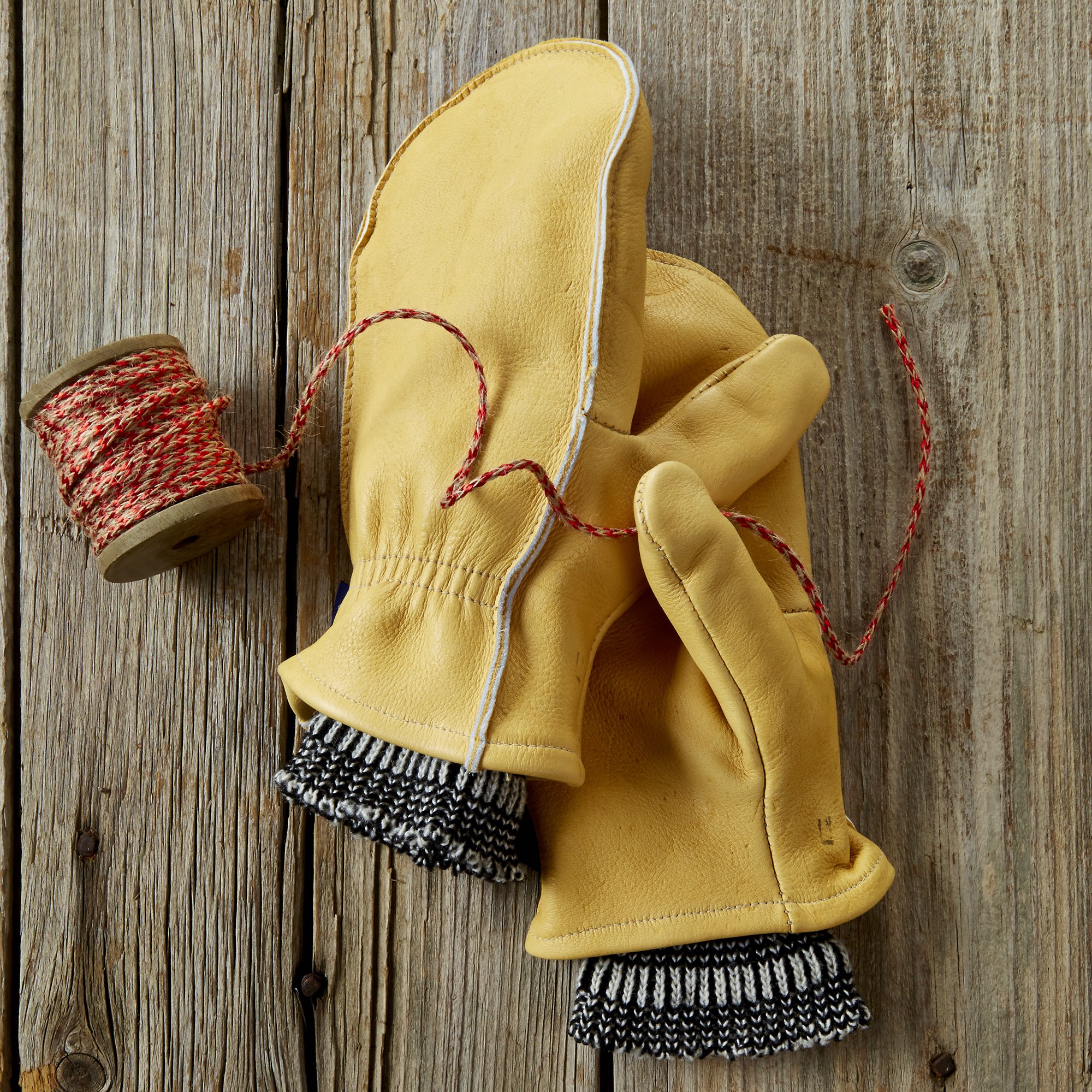 A pair of yellow work gloves with leather palms and cuffs lying on a wooden surface. Next to the gloves is a spool of red and white baker's twine and a piece of black and white knitted fabric.