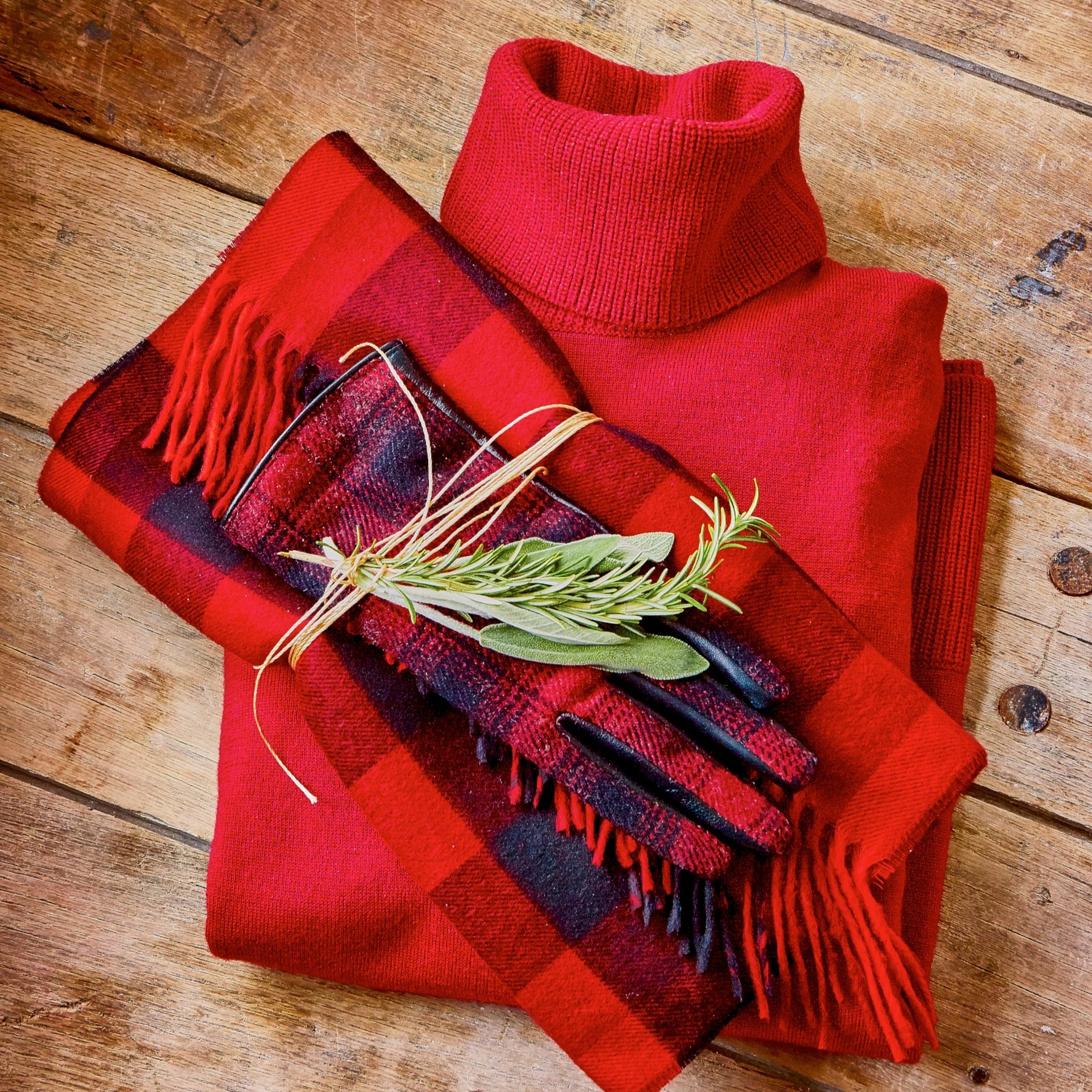 A red turtleneck sweater with fringed scarf, greenery, and string in a flat-lay on a wooden surface.