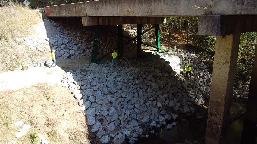 Construction workers in yellow safety vests working under a bridge, stabilizing the foundation with large rocks.