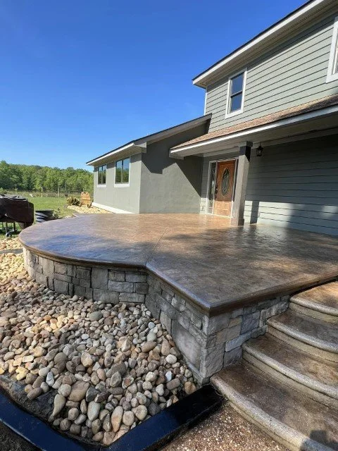Newly constructed house with a curved concrete patio and stone foundation, surrounded by rocks, on a sunny day.