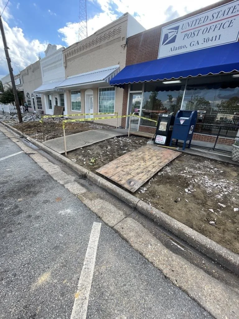 Sidewalk in front of a post office building under construction, with a temporary wooden ramp and caution tape, in Alamo, Georgia.