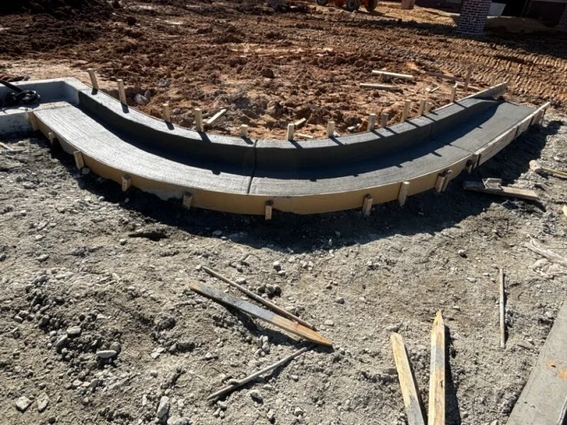 Curved concrete formwork and wooden supports on a construction site, with exposed dirt and scattered building materials.