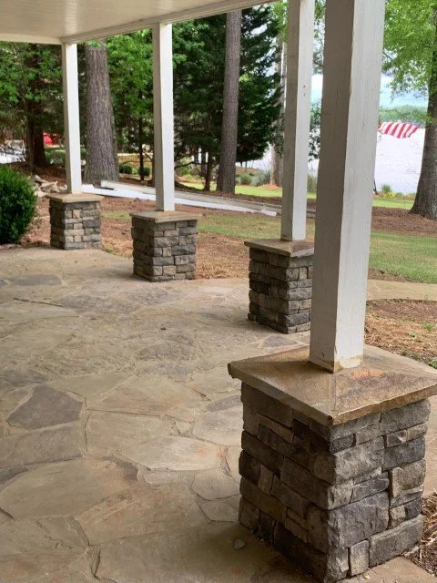 Stone and wood porch with four brick pillars supporting a roof, overlooking a yard with trees and a white building in the background.