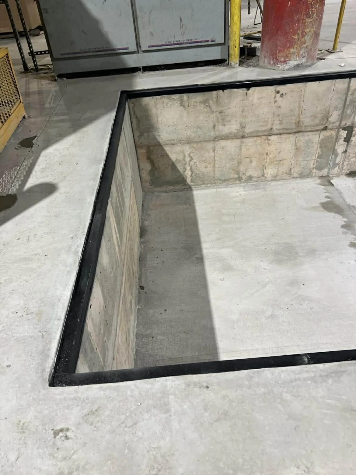 Empty concrete pit on construction site, framed with metal, with surrounding concrete floor and construction materials in background.