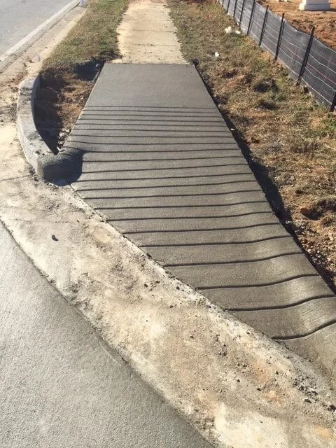 Newly poured concrete sidewalk next to a dirt and grass area, with a black mesh fence on the right side.