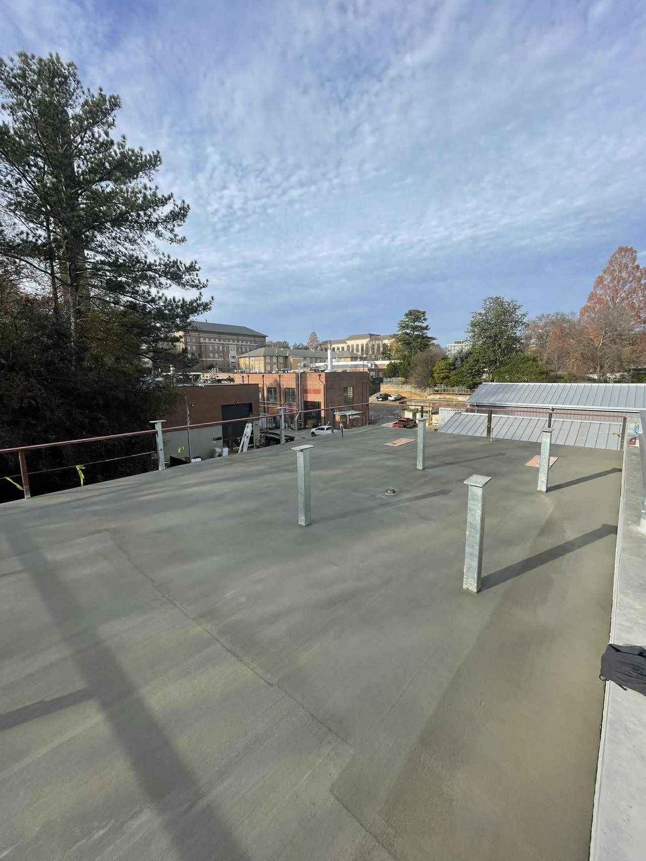 View of a rooftop with metal post supports and a safety railing in an urban setting during the day, with trees and buildings in the background under a partly cloudy sky.