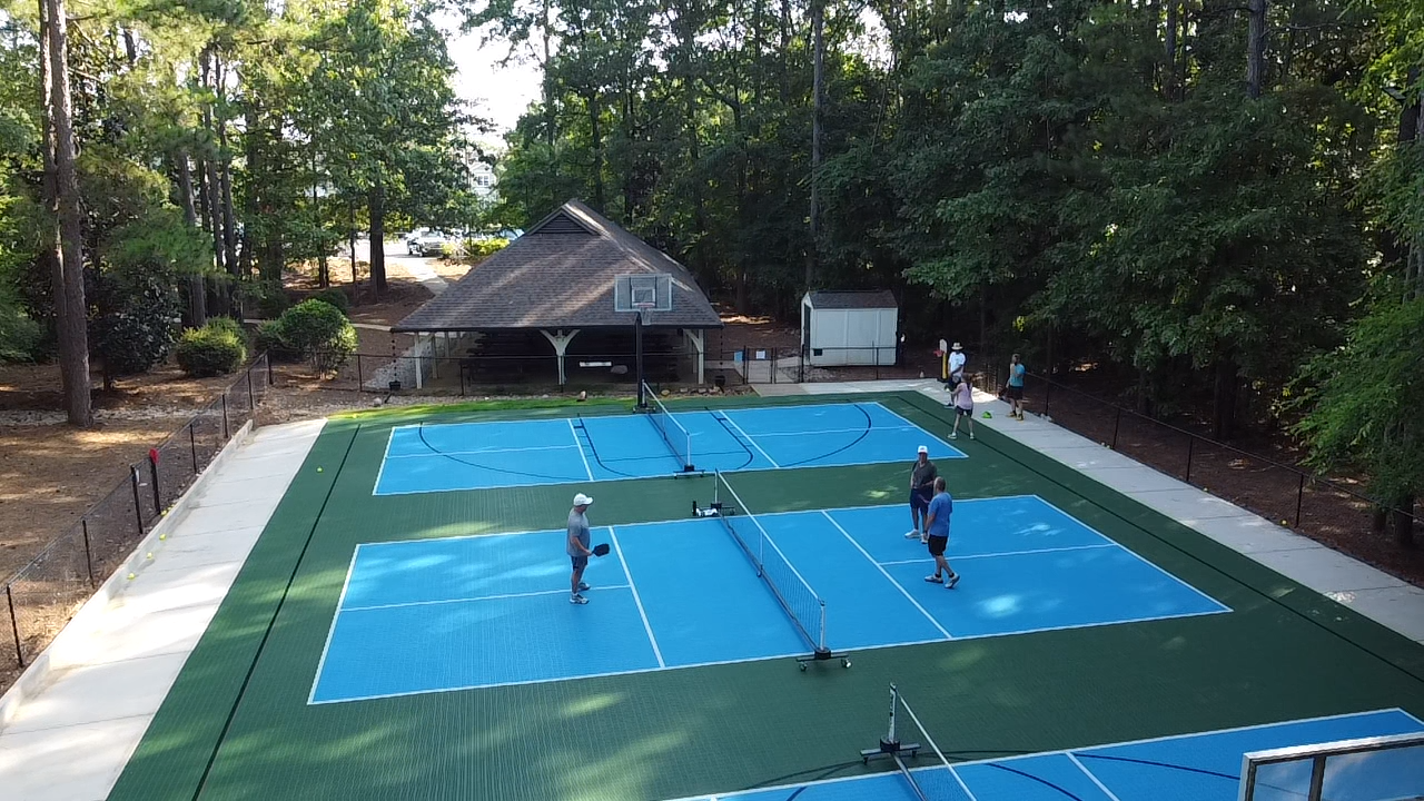 People playing pickleball on a blue and green court surrounded by trees, with a basketball hoop and a small building or shed in the background.