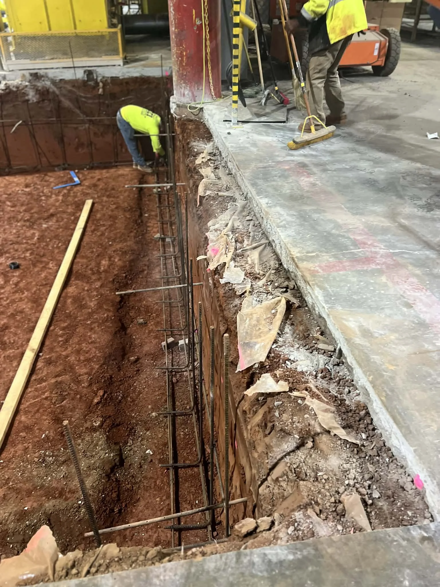 Construction workers are working on a construction site. One worker is inside a dug hole, and another is standing on a concrete surface with construction tools nearby. The dug hole has exposed rebar and dirt, indicating ongoing foundational work.