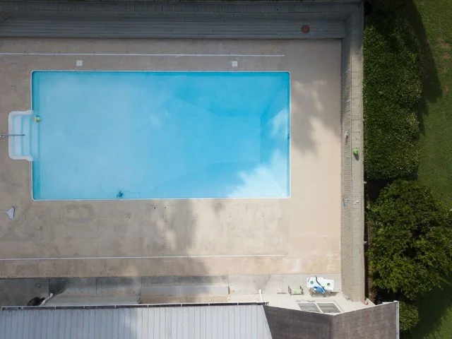 An aerial view of a rectangular in-ground swimming pool with blue water, surrounded by a concrete deck and green trees.