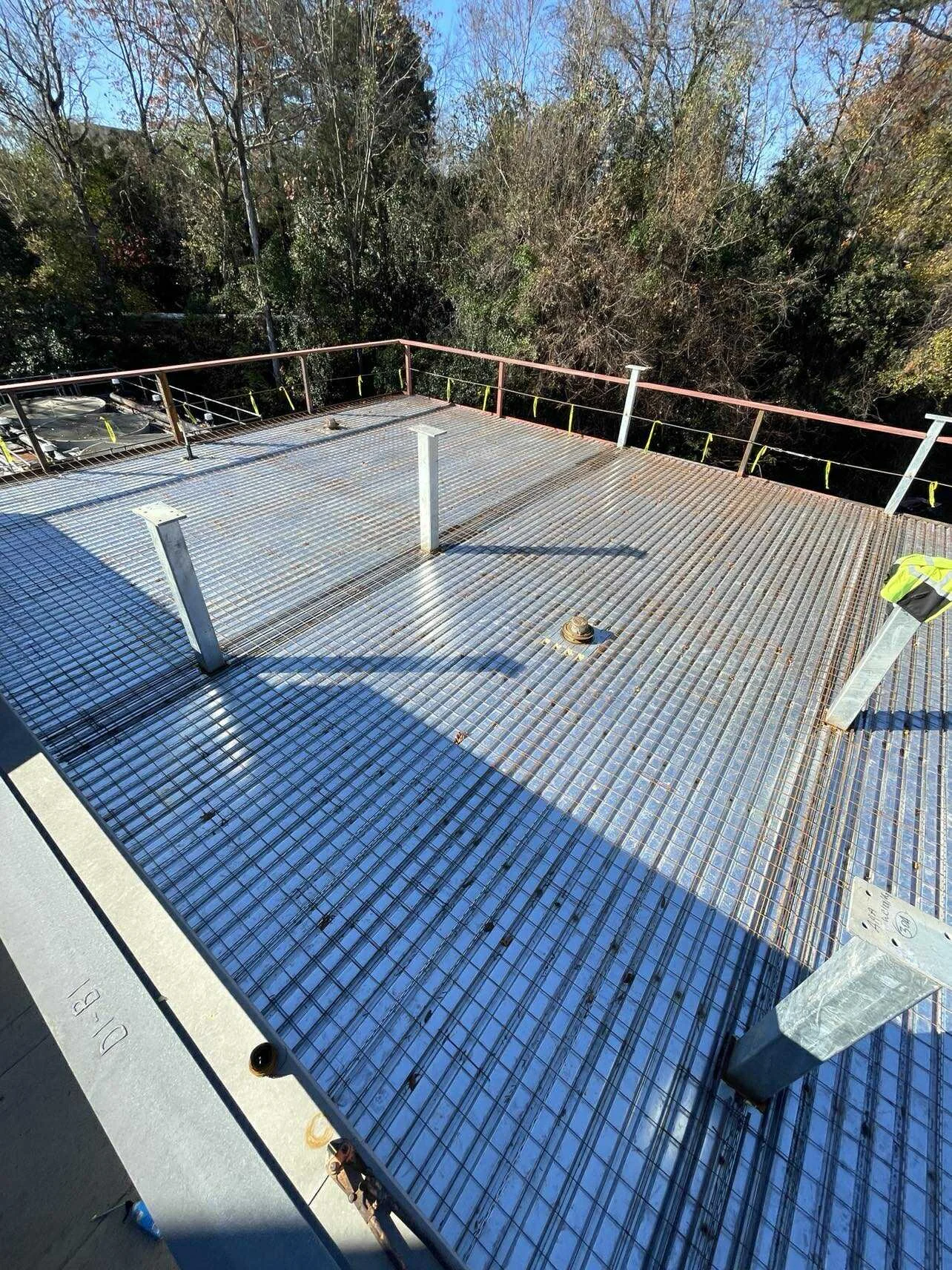 Construction site on the roof of a building with a metal grid and ventilation pipes, surrounded by safety railings and trees in the background.
