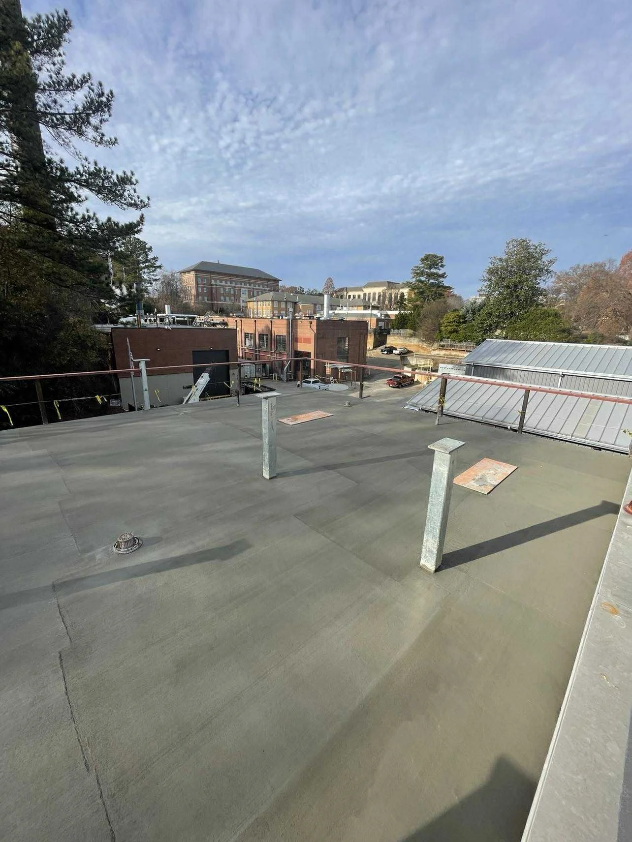View of a rooftop with a concrete surface, some vent pipes, and a safety railing. In the background, there are buildings and trees under a partly cloudy sky.