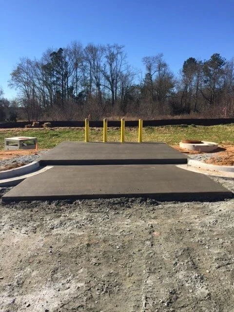 A newly paved parking lot with two levels, separated by a curb, and yellow safety posts at the top level, with trees and a clear blue sky in the background.