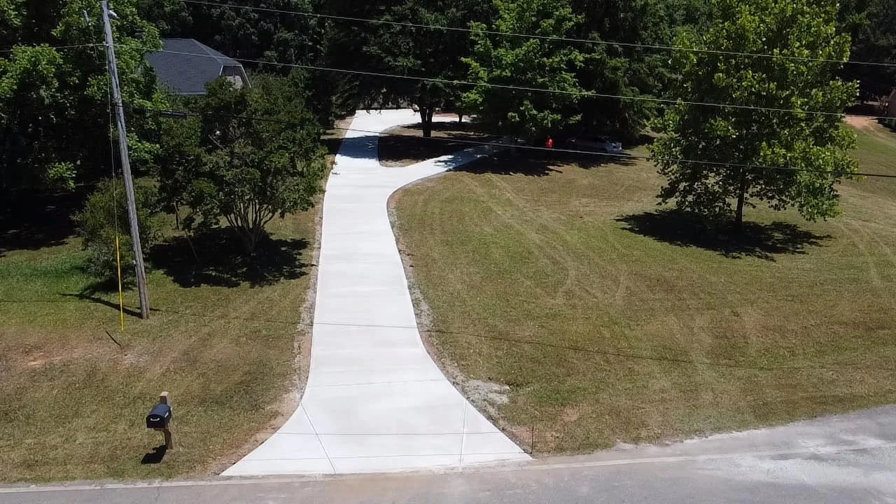 A curved concrete driveway leading to a house, with grass and trees on either side and a mailbox near the street.