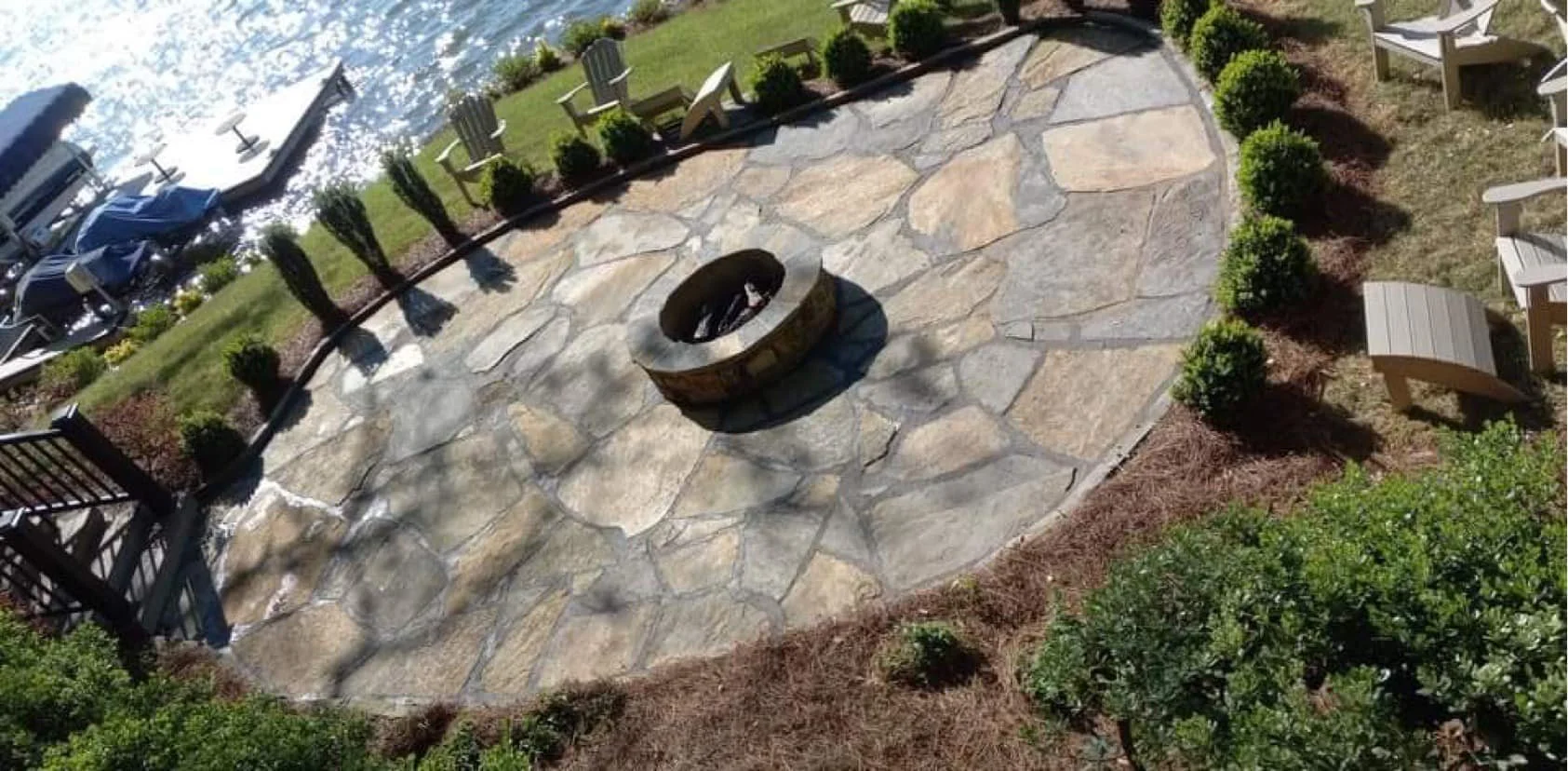 A stone fire pit circle on a stone patio surrounded by bushes and potted plants, with Adirondack chairs and a walkway nearby, overlooking a body of water with boats docked.