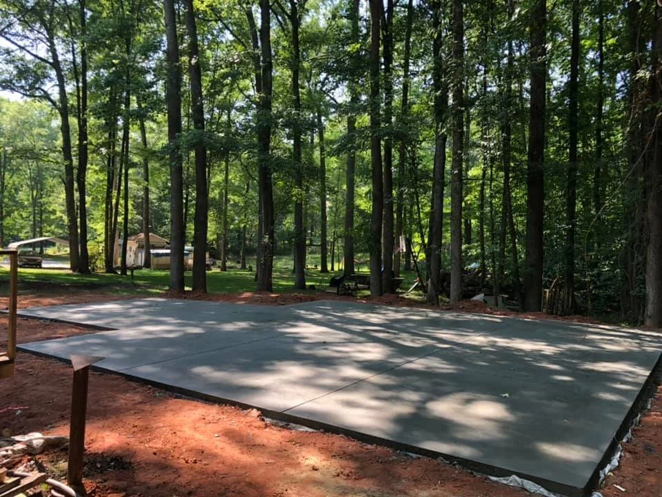 Concrete foundation laid on red dirt in a wooded area with tall trees, supporting a construction site.