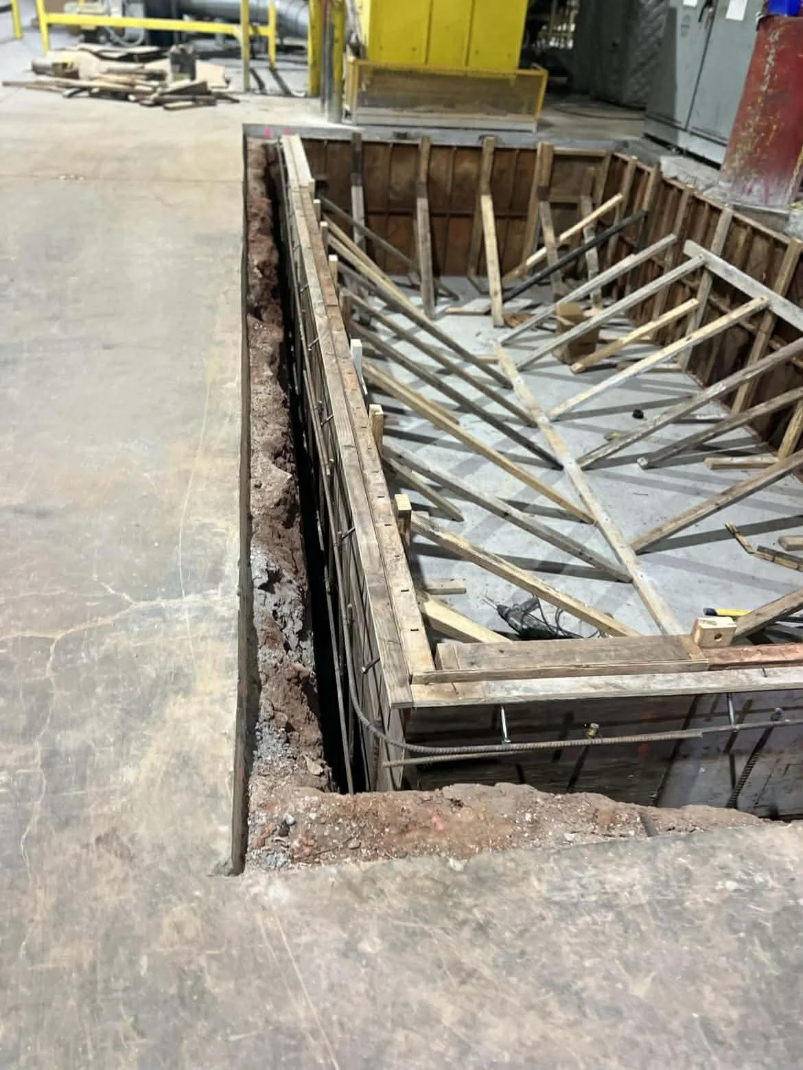 Construction site showing a square hole in the ground framed with wood, with various wooden supports and concrete bottom.