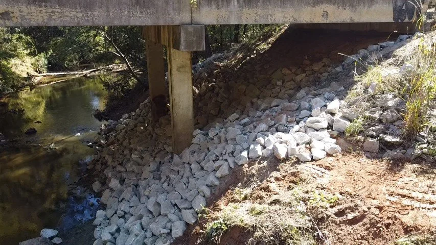 Storm drain near water with gravel and soil, under a concrete bridge or road, surrounded by trees and vegetation.
