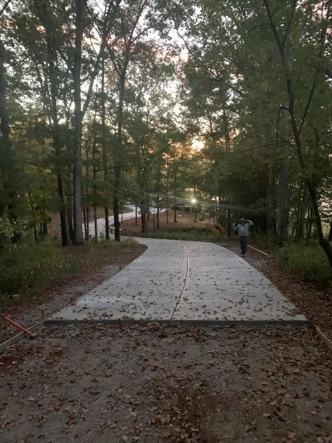 A winding concrete sidewalk through a wooded park area during sunset, with a person walking away from the camera.
