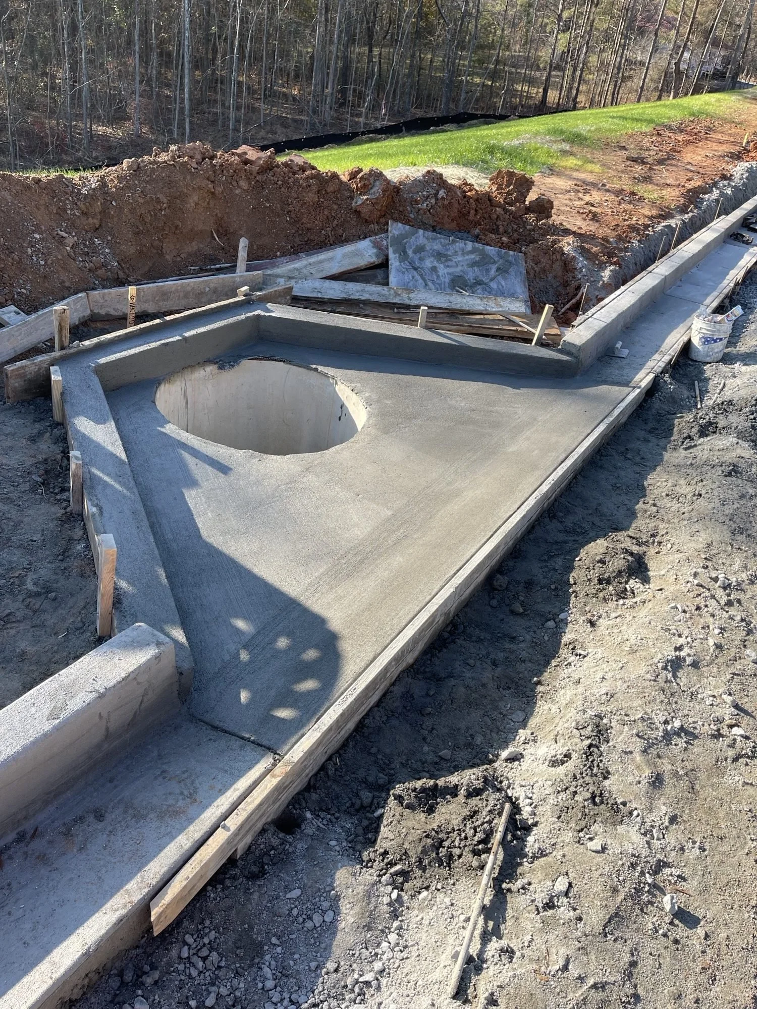 Under construction sidewalk with concrete and wooden forms, dirt, and gravel, with green grass and trees in the background.