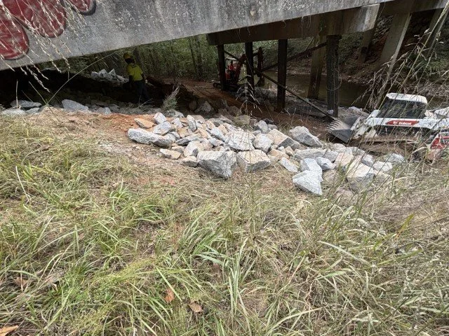 A collapsed bridge with support beams damaged, rocks and debris scattered below, and emergency personnel on site.